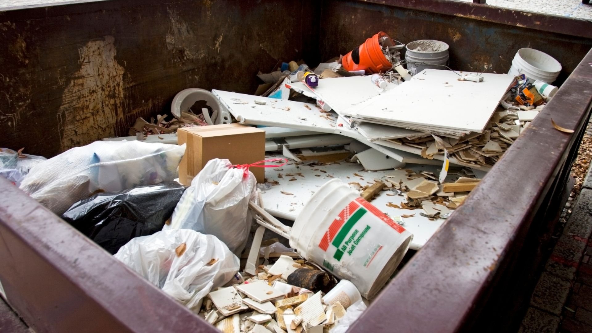 Overflowing dumpster filled with construction debris, bags, and broken materials