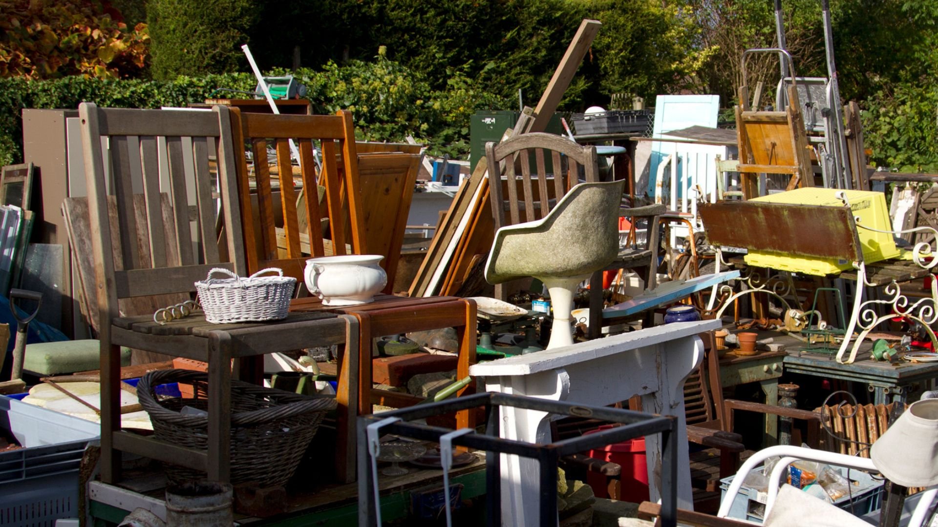 Cluttered yard sale with various furniture, chairs, baskets, and household items
