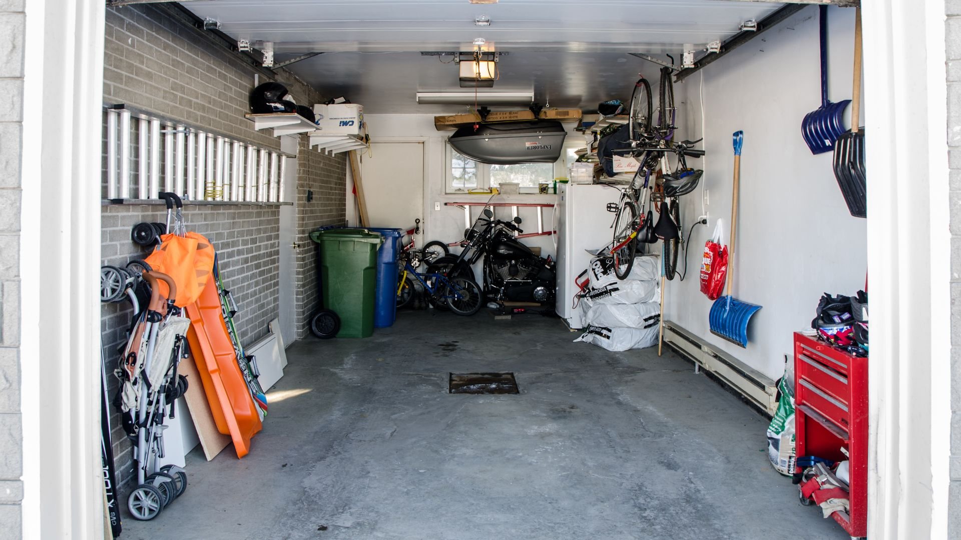 Cluttered garage interior with bikes, tools, snow shovels, and storage containers